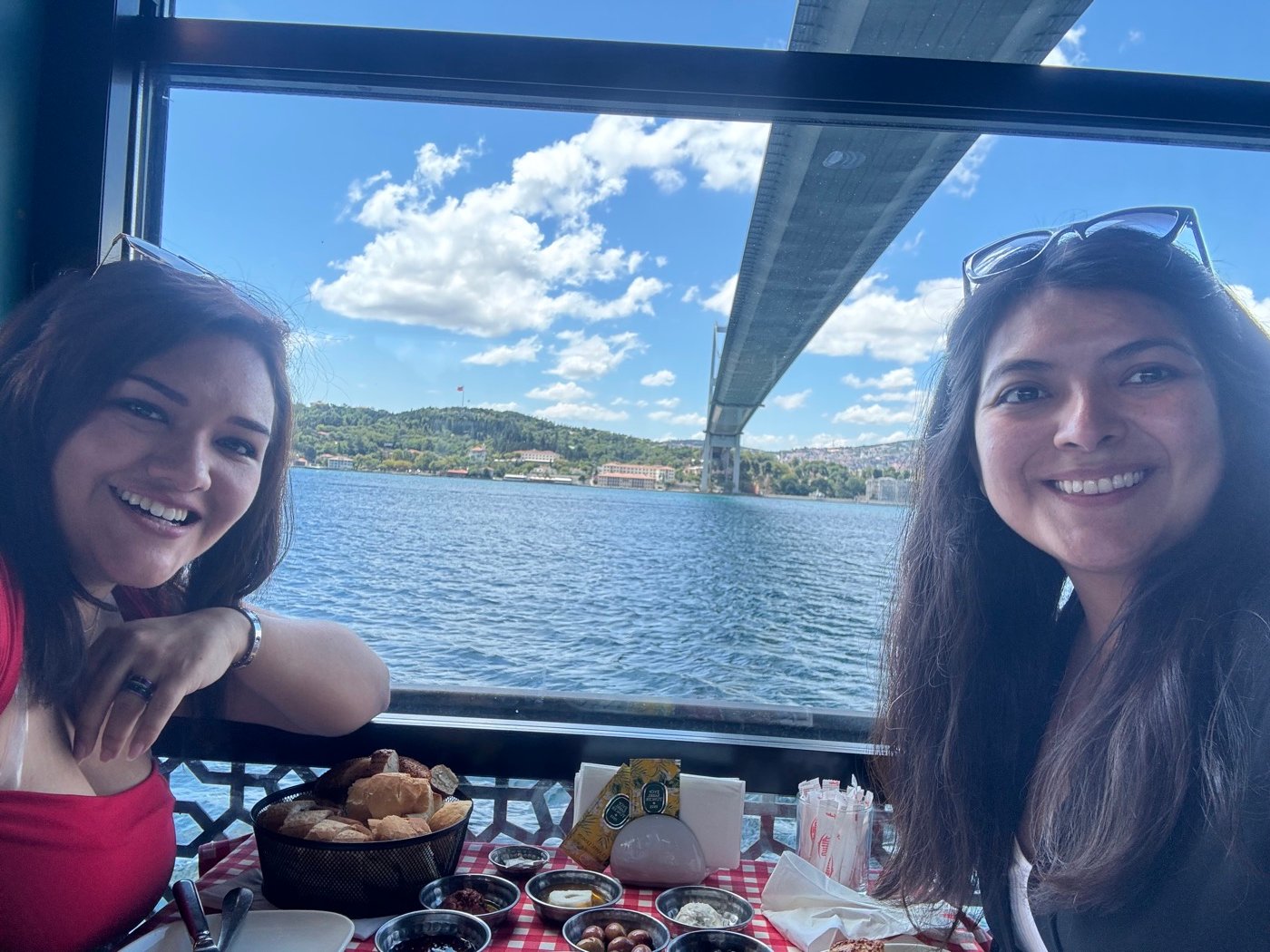 Mariel Cárdenas y una amiga sonriendo durante el desayuno, con el puente del Bósforo de fondo